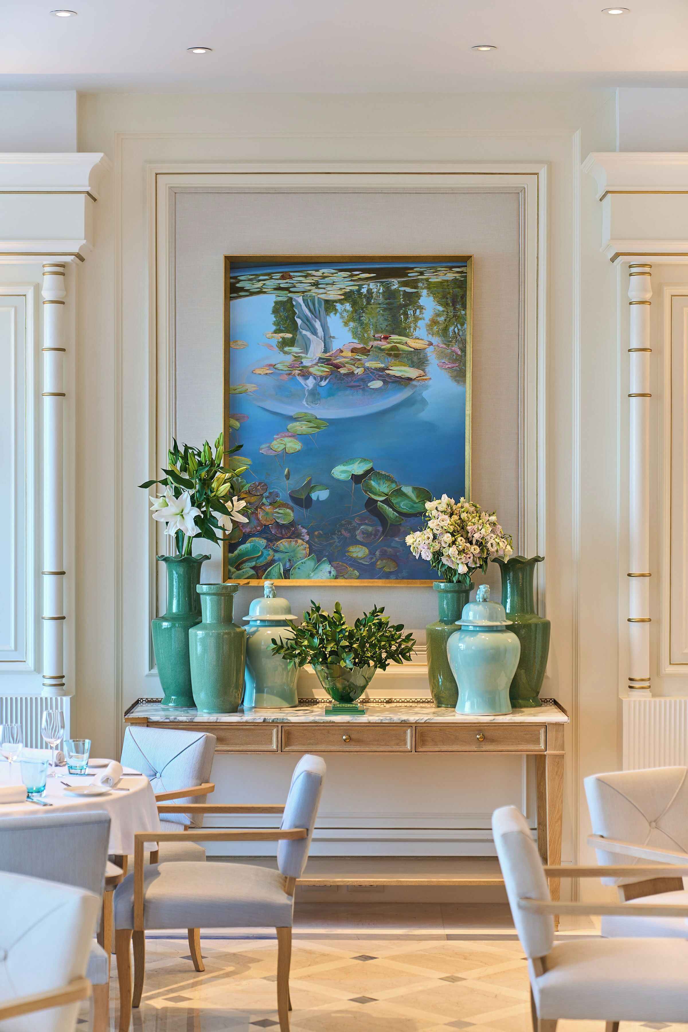 A dining area with a large painting of a pond and flowers, green vases, and white chairs.