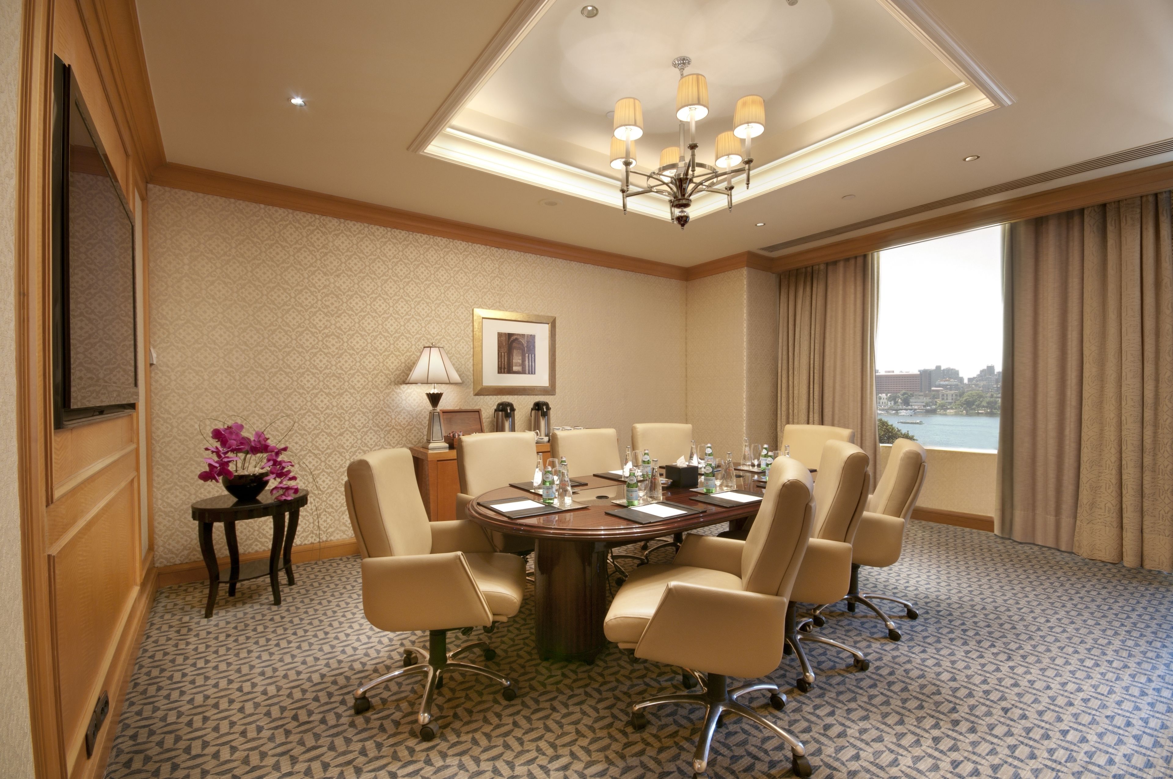 A conference room with a round table, chairs, a chandelier, a lamp, a framed picture, and a view of a cityscape through large windows.