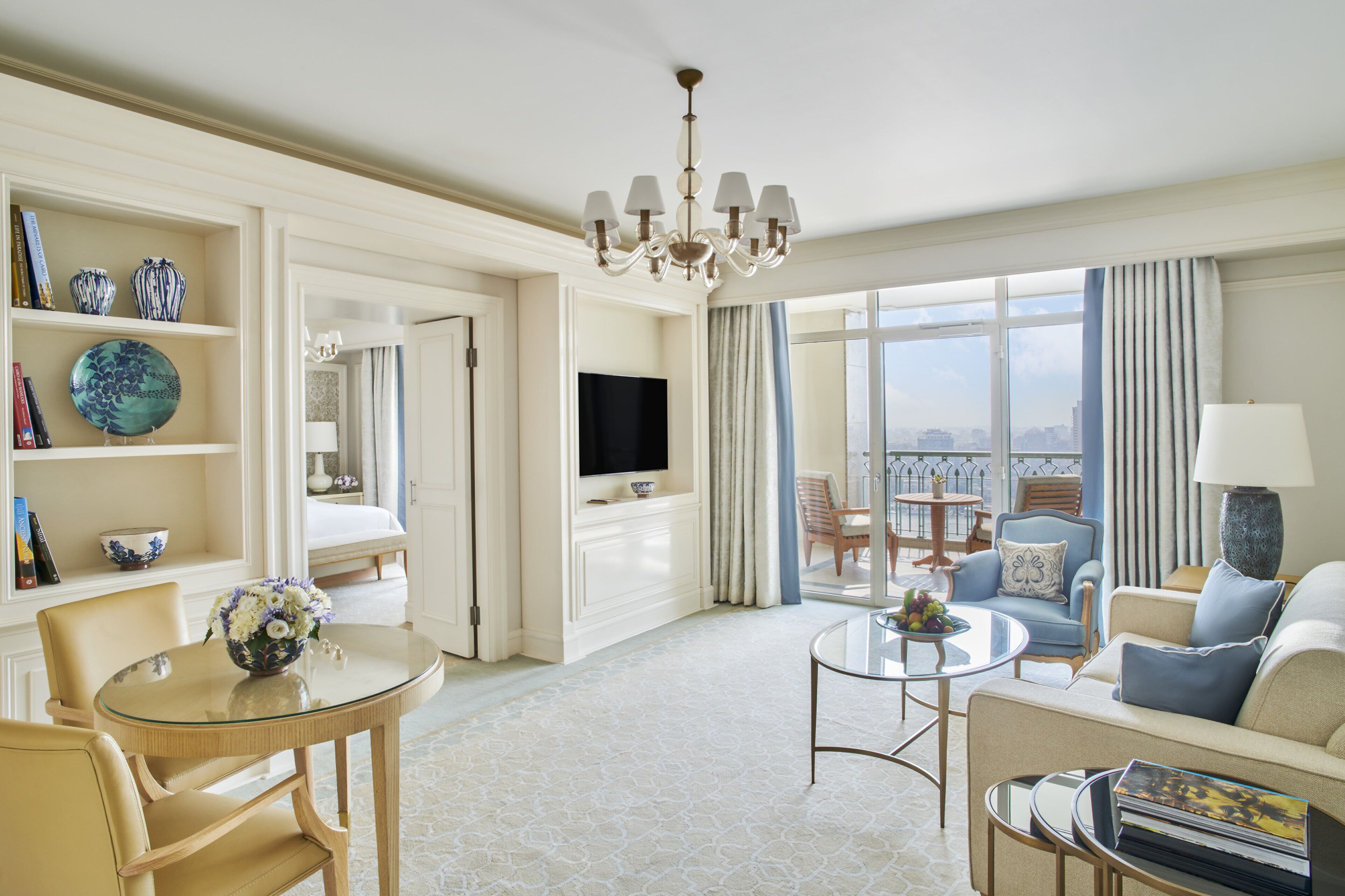 A spacious hotel room with a balcony, a television, a dining area with a glass table, and a seating area with a sofa and armchairs.