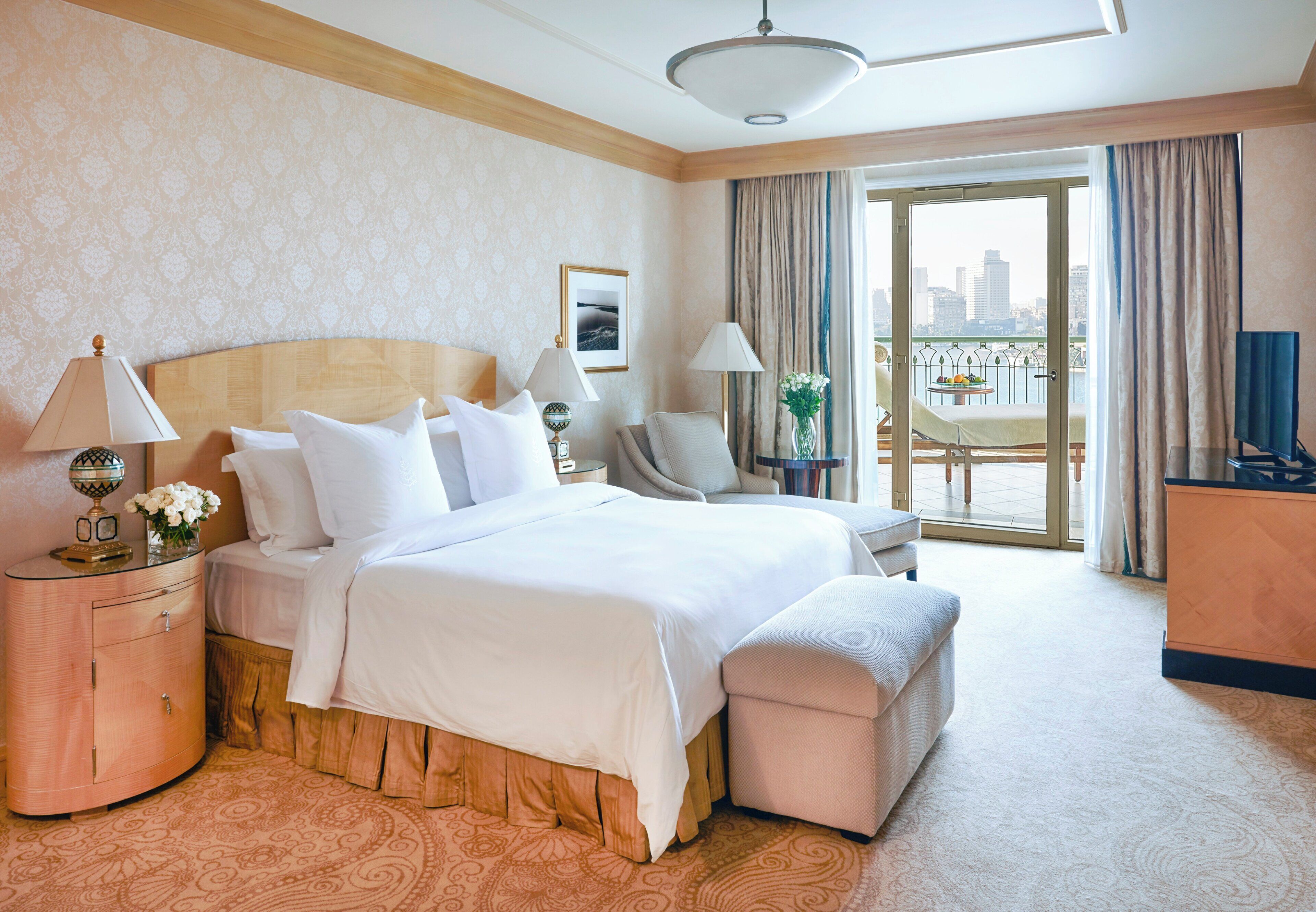 A hotel room with a large bed, a desk, a chair, a balcony with a view of the city, and a television.