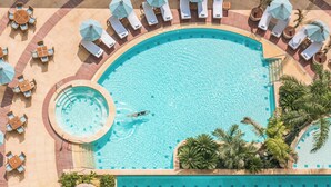 Vue aérienne d’une piscine entourée de chaises longues et de parasols, avec des palmiers et un jacuzzi circulaire à proximité.