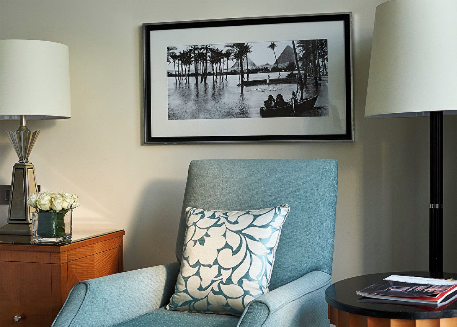A room with a framed black and white photograph of a water scene with palm trees and pyramids, a blue armchair with a patterned pillow, a side table with a lamp and a vase of flowers, and a small round table with a magazine.