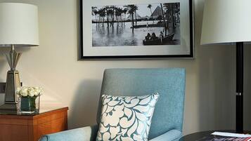 A room with a framed black and white photograph of a water scene with palm trees and pyramids, a blue armchair with a patterned pillow, a side table with a lamp and a vase of flowers, and a small round table with a magazine.