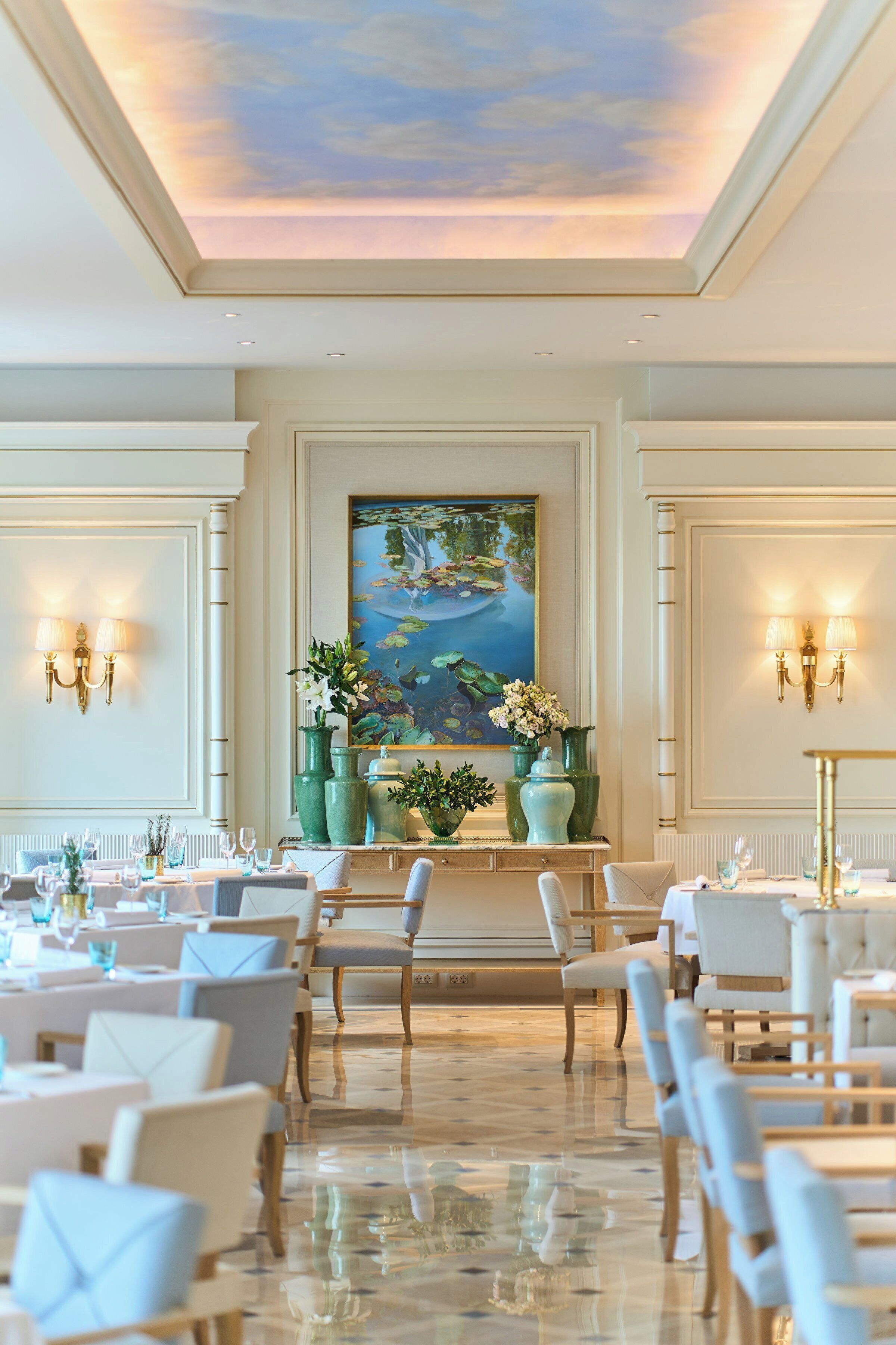 Un espace repas avec des tables dressées pour le repas, des chaises, un plafond décoré d’un ciel peint et des luminaires fixés au mur.