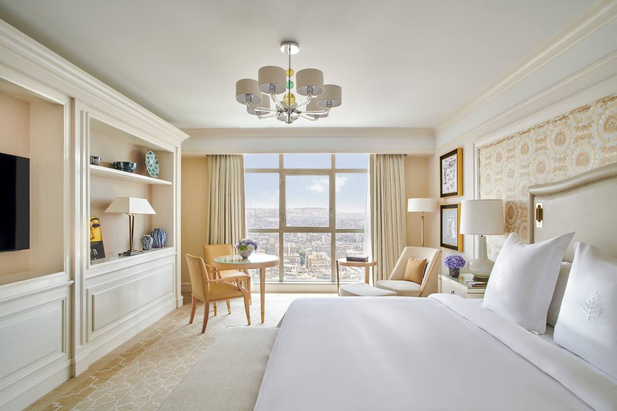 Ein Hotelzimmer mit einem großen Bett, einer Kristallleuchter, einem Schreibtisch mit Stuhl, einem Fernseher und Blick auf die Stadt durch große Fenster.