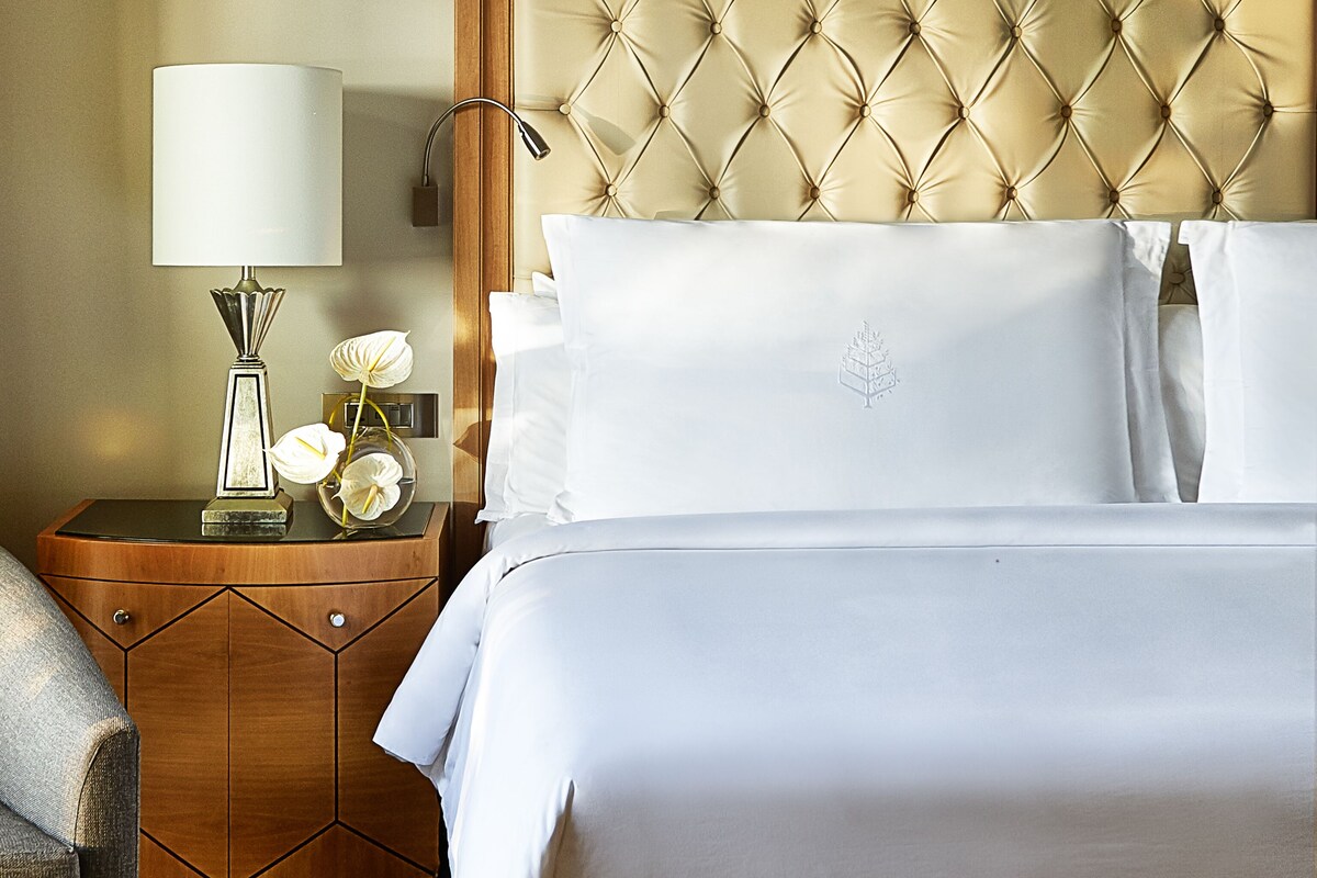 Une chambre d’hôtel avec une tête de lit capitonnée, une table de chevet avec une lampe et des fleurs, et un lit parfaitement fait.
