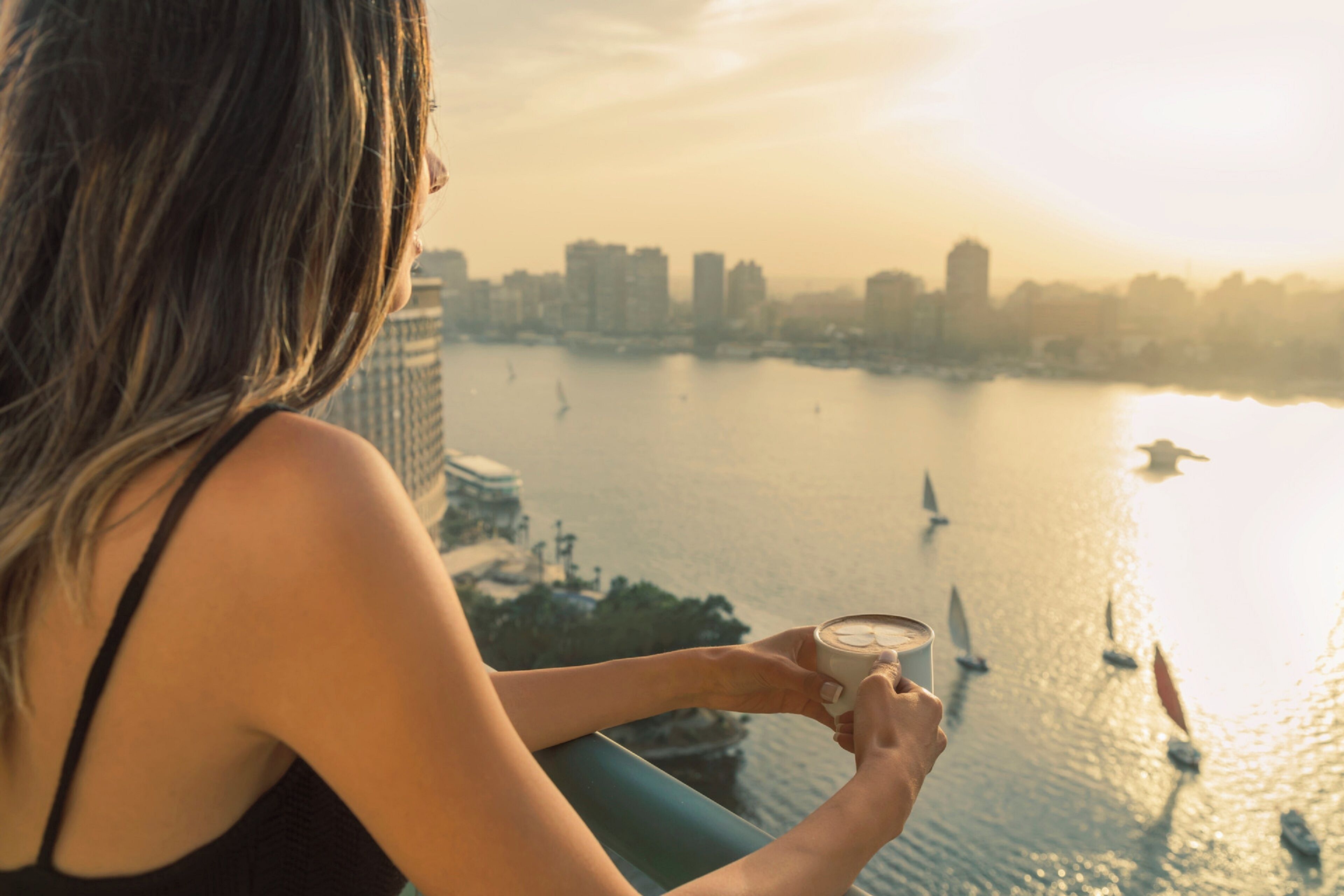 Una persona sosteniendo una taza, con vista a un cuerpo de agua con veleros y edificios al fondo.
