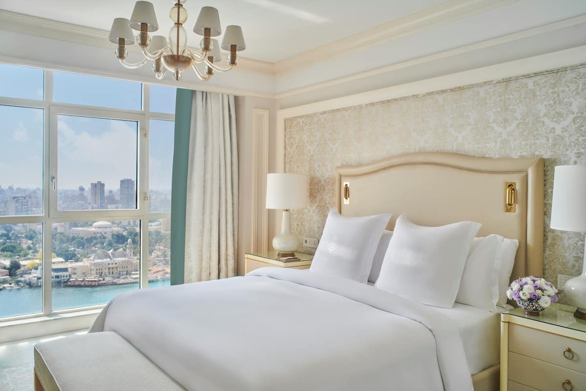 Ein Hotelzimmer mit einem großen Bett, einer Kristallleuchter, Blick auf die Stadt und einem Nachttisch mit Lampe und Blumen.
