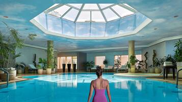 Una amplia piscina interior con techo de tragaluces, rodeada de plantas en macetas y sillas de descanso.