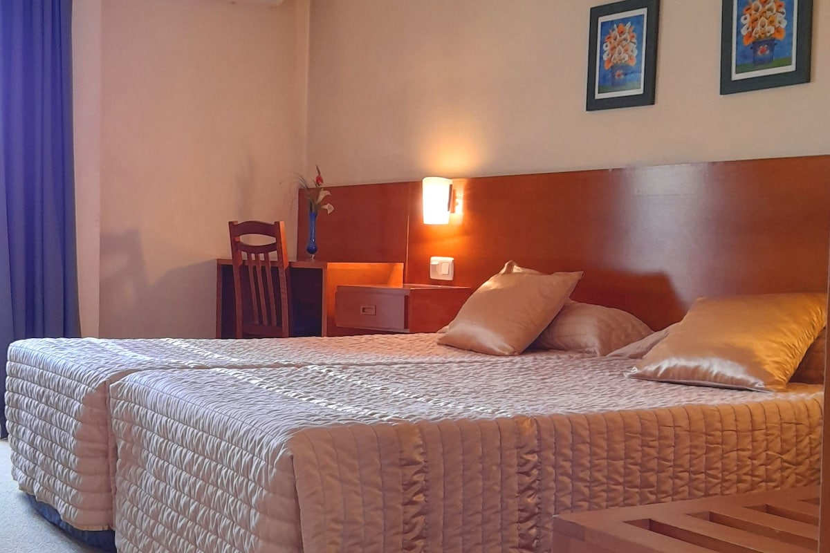 A hotel room with two beds, a desk, a chair, an air conditioning unit, and two framed pictures on the wall.