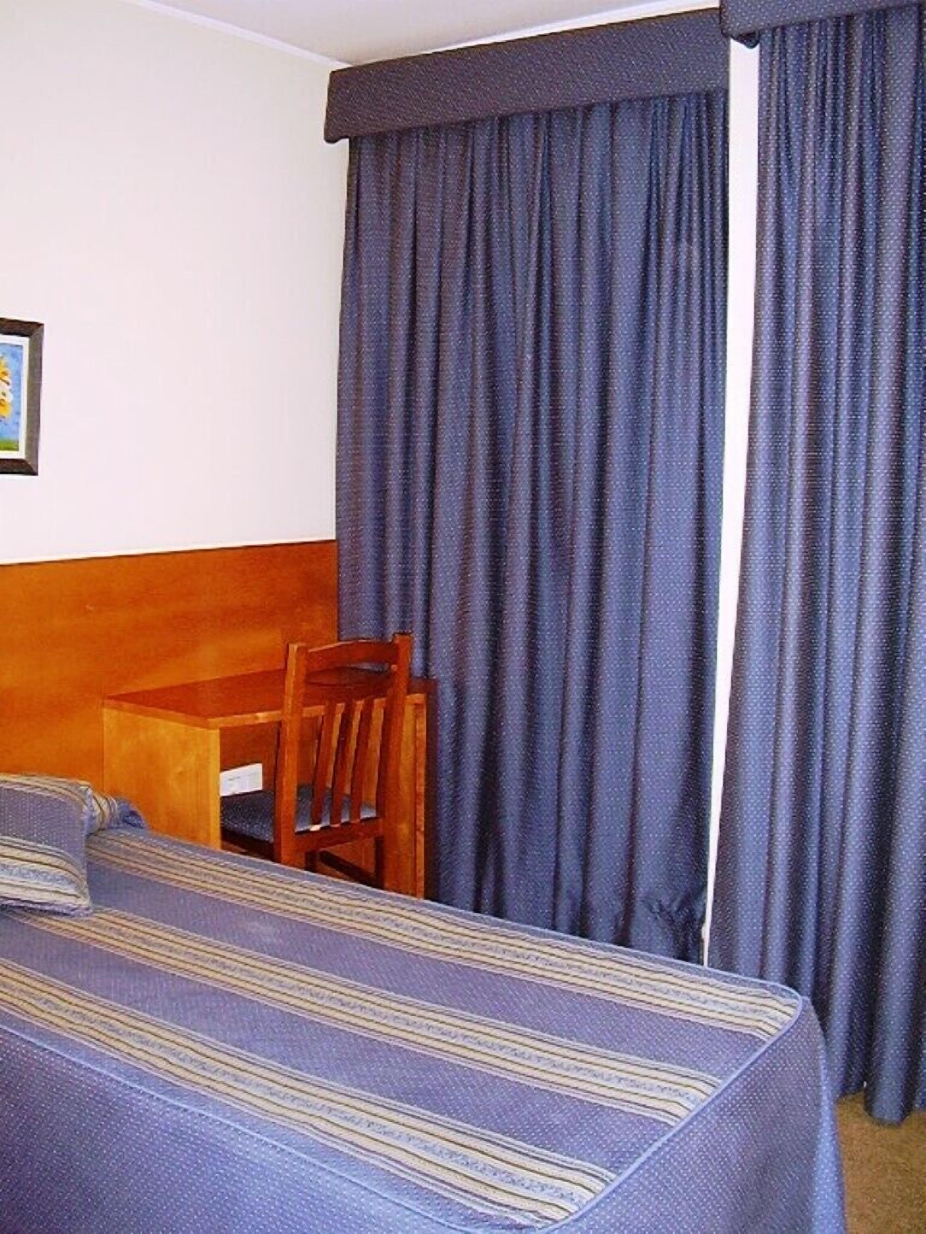 A hotel room with a bed, a wooden chair, and a window with dark curtains.