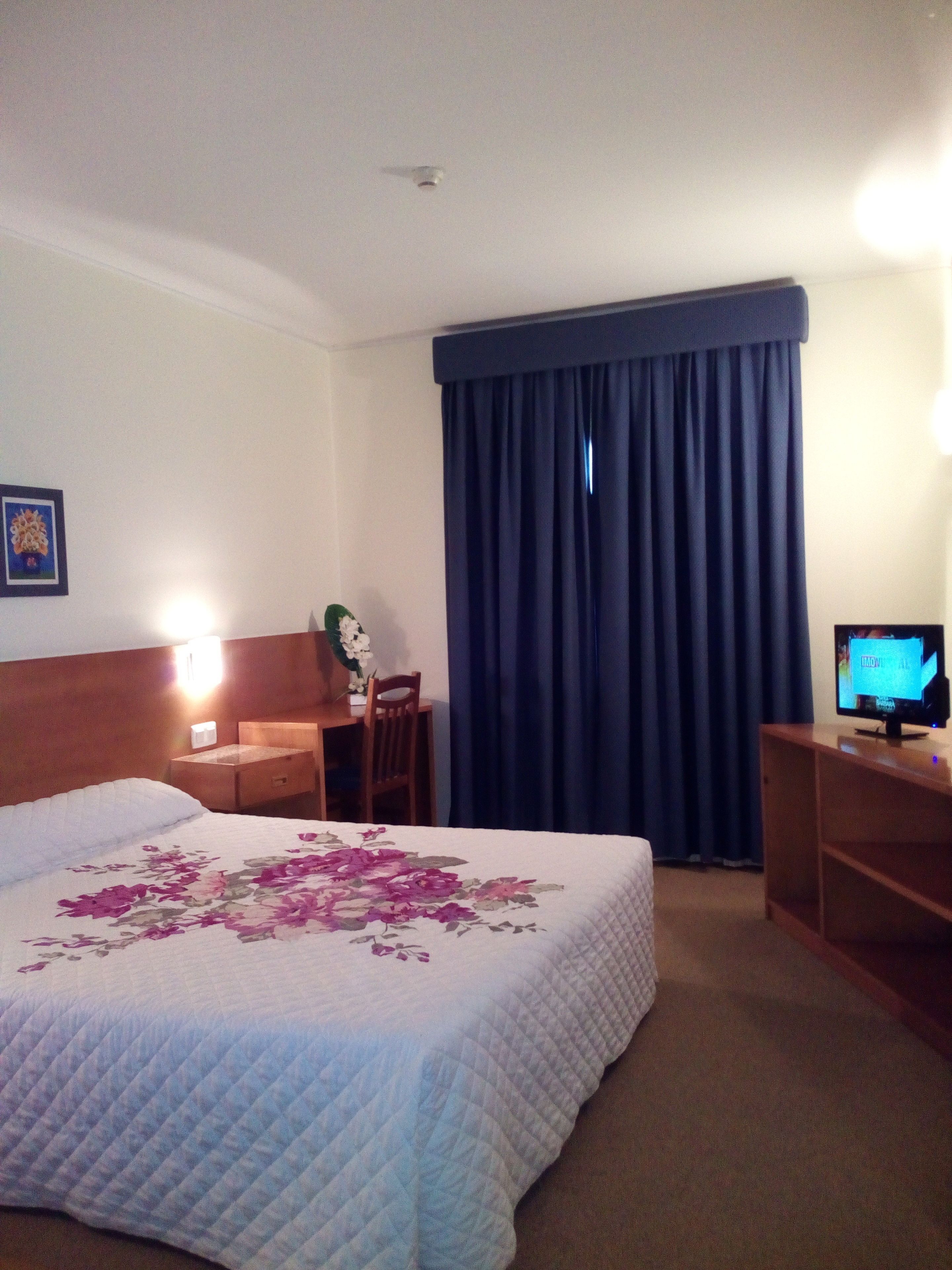 Habitación de hotel con cama, escritorio, silla, televisor y ventana con cortinas.
