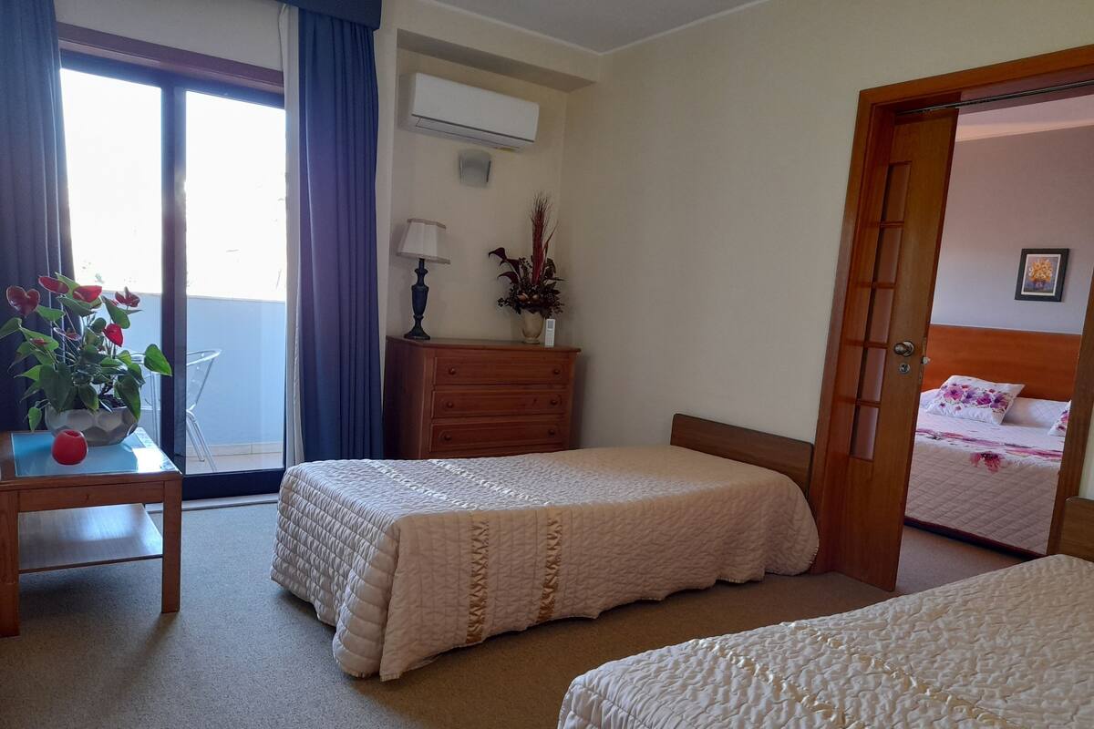 A hotel room with two beds, a wooden dresser, a desk with a vase, and a window with curtains.