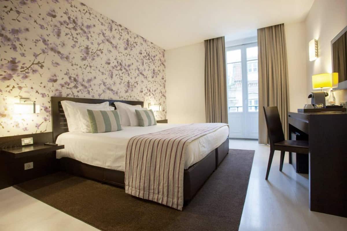 A hotel room with a large bed, a desk with a chair, a nightstand, and a window with curtains.