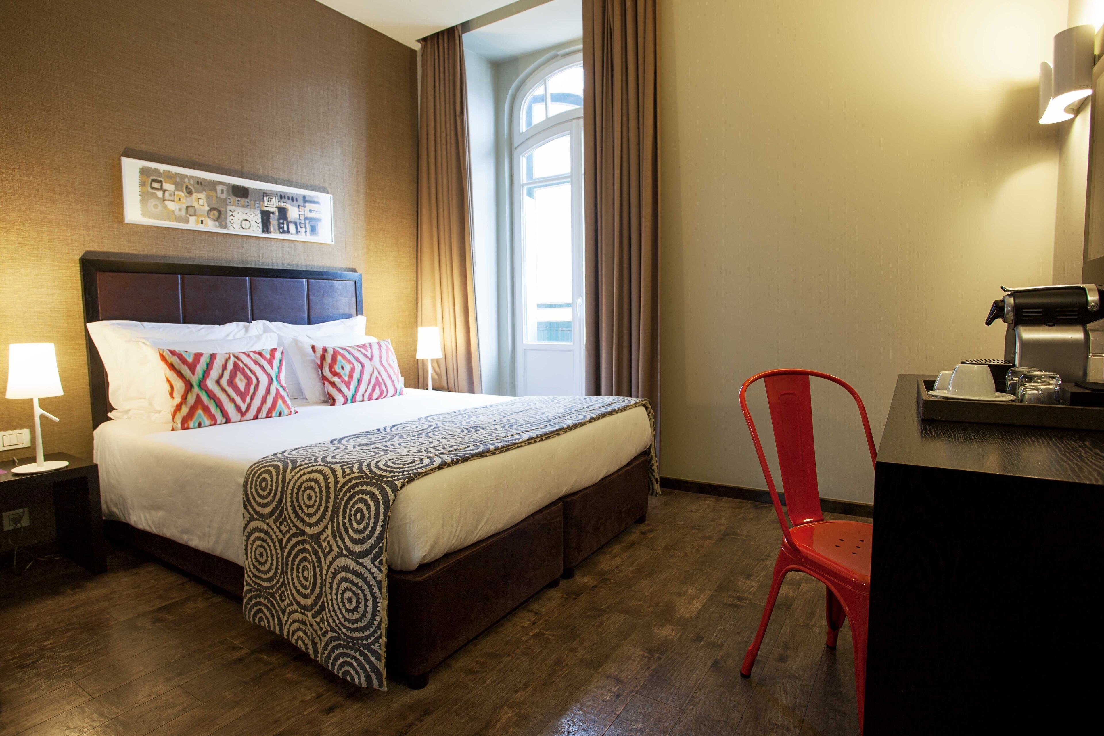 Habitación de hotel con una cama grande, una silla roja, un escritorio con cafetera y una ventana con cortinas.