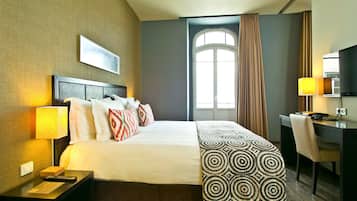 Ein Hotelzimmer mit einem großen Bett, einem Schreibtisch, einem Stuhl, einer Lampe und einem Vorhangfenster.