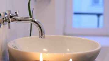 Um banheiro com uma pia branca, duas velas e uma flor roxa ao fundo.