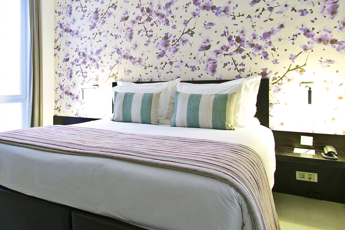 A neatly made bed with striped bedding, a floral wallpaper, and a bedside table with a lamp and phone.