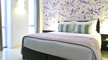 Una cama bien hecha con ropa de cama a rayas, un papel tapiz floral y una mesita de noche con una lámpara y un teléfono.