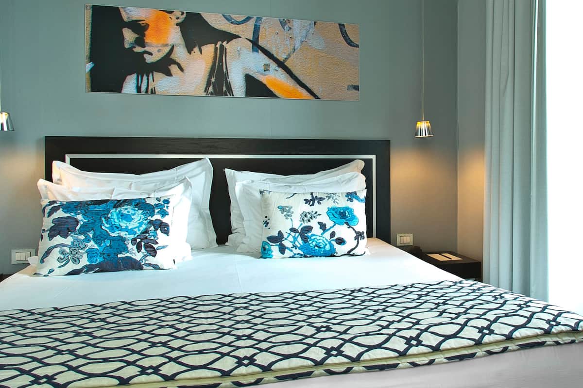 A hotel room with a large bed, two pillows with floral patterns, a geometric-patterned bedspread, a framed abstract painting above the bed, and a window with curtains.