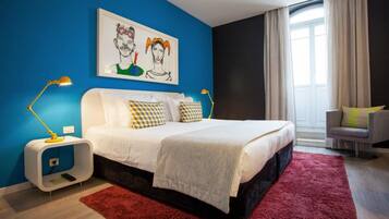 Un dormitorio moderno con una pared azul decorativa, una cama grande con almohadones estampados, una alfombra roja y una mesita de noche con lámpara.