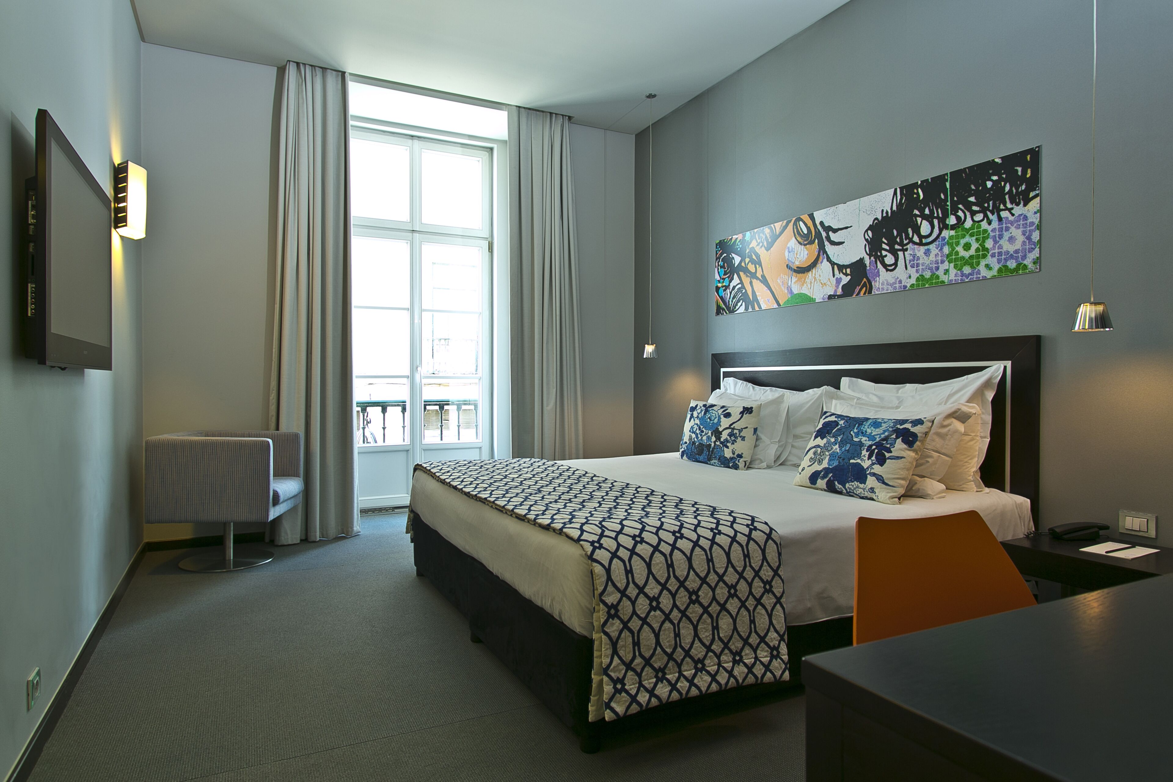 A modern hotel room with a large bed, a desk, a chair, a radiator, and a large window with curtains.
