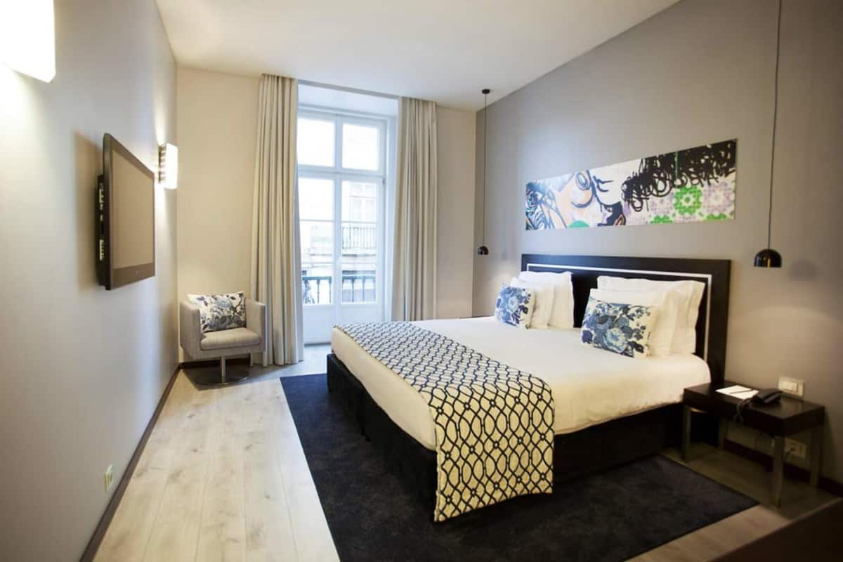 A modern hotel room with a large bed, a chair, a desk, and a view of the outdoors through a window.