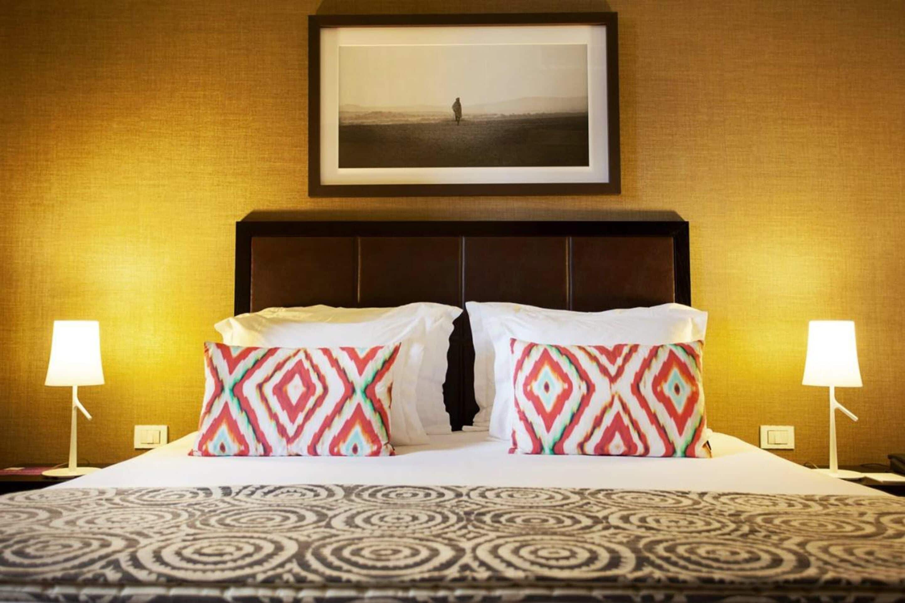 Quarto de hotel com uma cama grande, duas almofadas estampadas, um quadro na parede e duas luminárias de cabeceira.