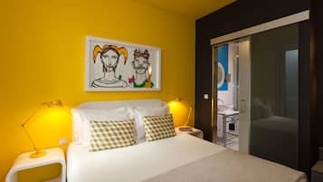 A modern hotel room with a large bed, two bedside tables with lamps, a painting on the wall, and a bathroom visible through a glass door.