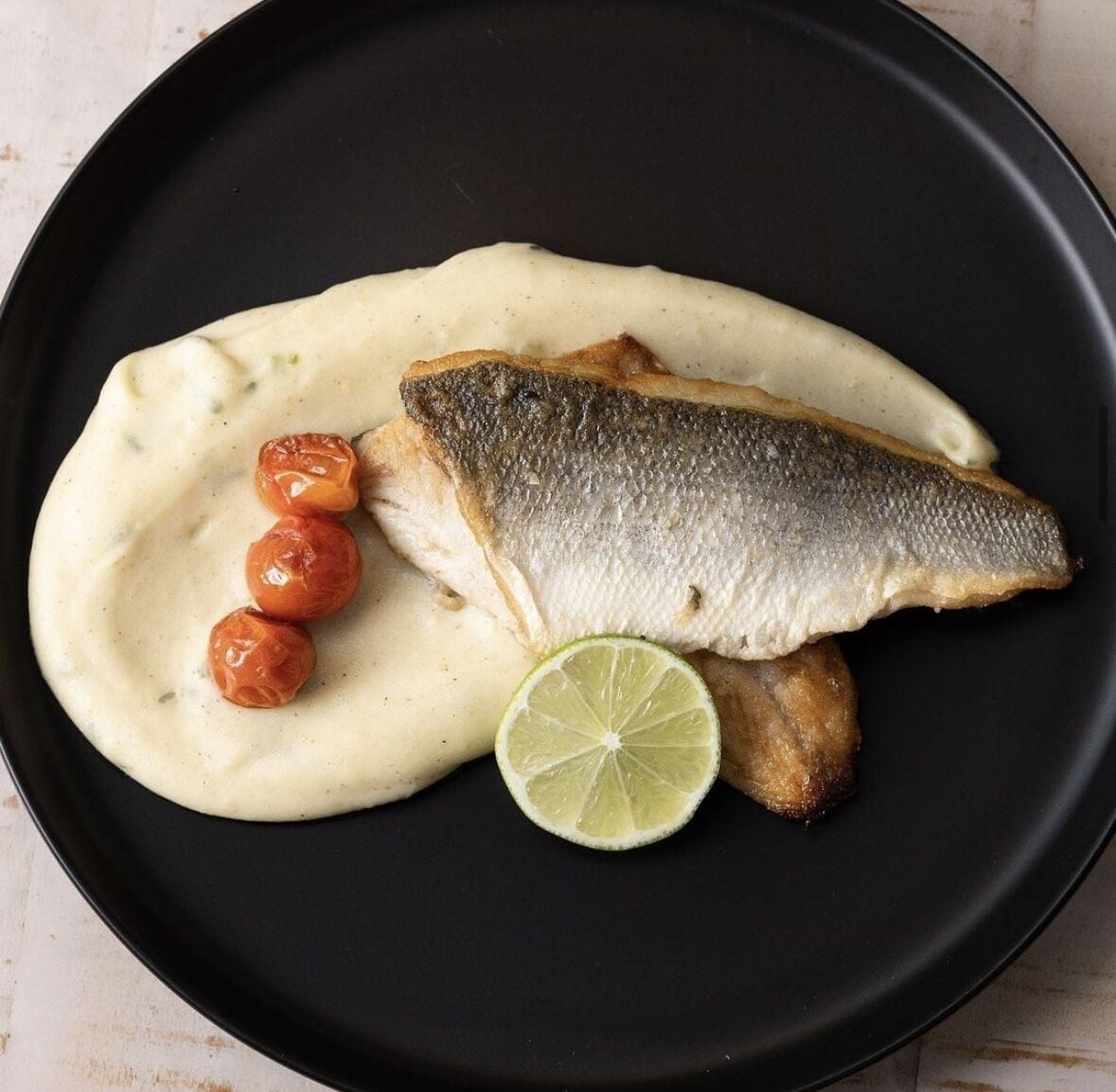 Une assiette noire avec un poisson, une sauce crémeuse, des tomates cerises et une tranche de citron vert.