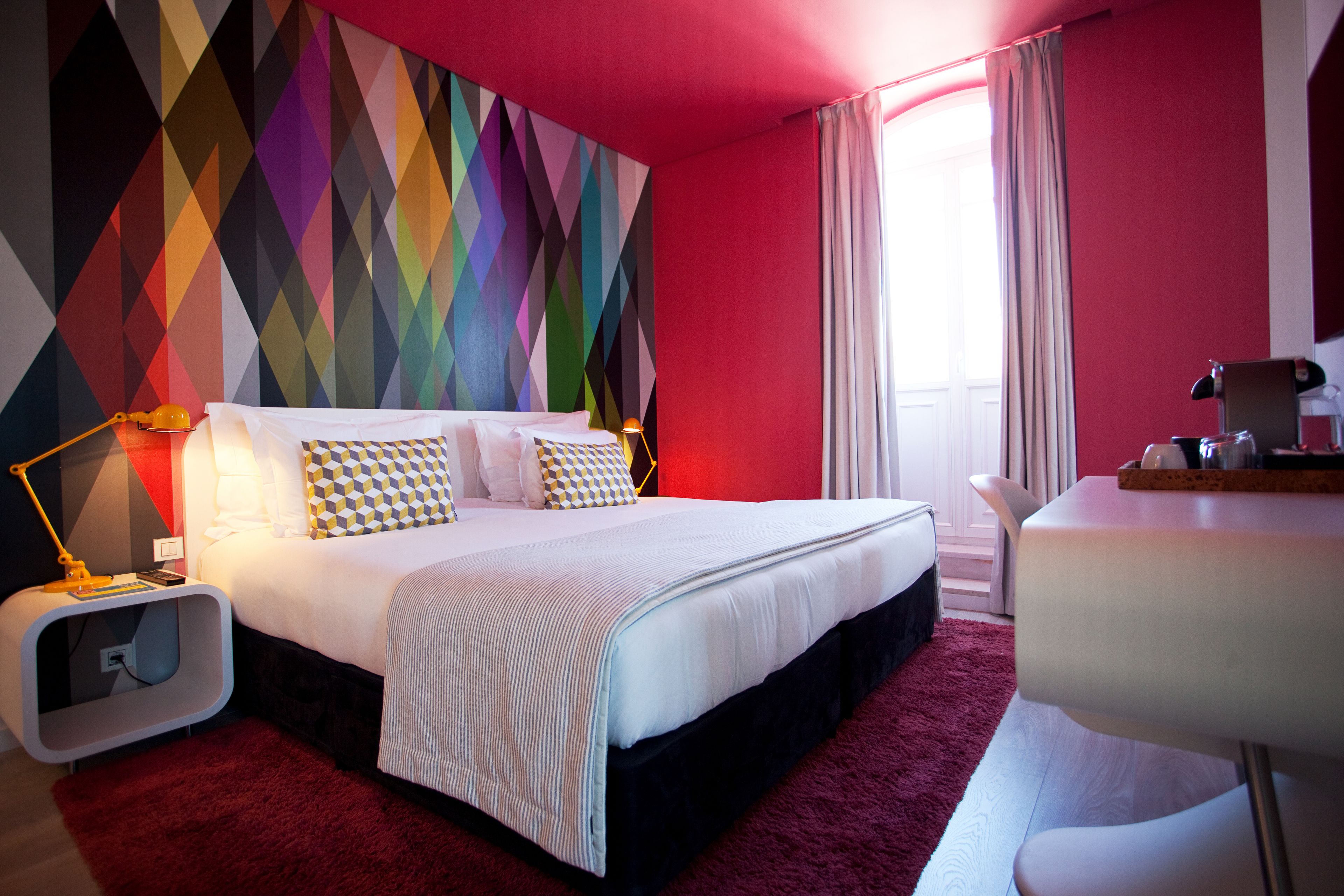 A modern hotel room with a large bed, a desk, a colorful geometric-patterned wall, and a red carpet.