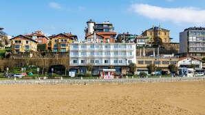 Un hotel frente a la playa con varios pisos, una playa arenosa al frente y diversos edificios en el fondo.