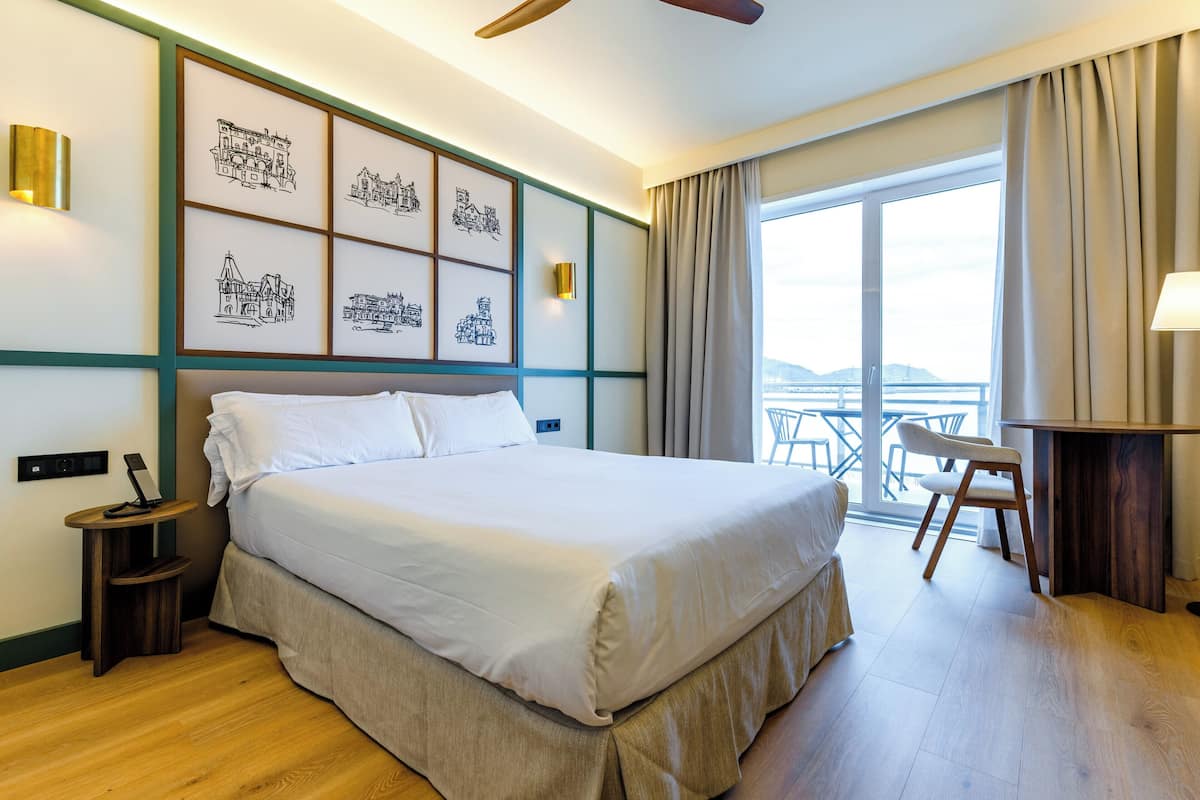 A hotel room with a large bed, a desk, a chair, a balcony with a view, and artwork on the wall.
