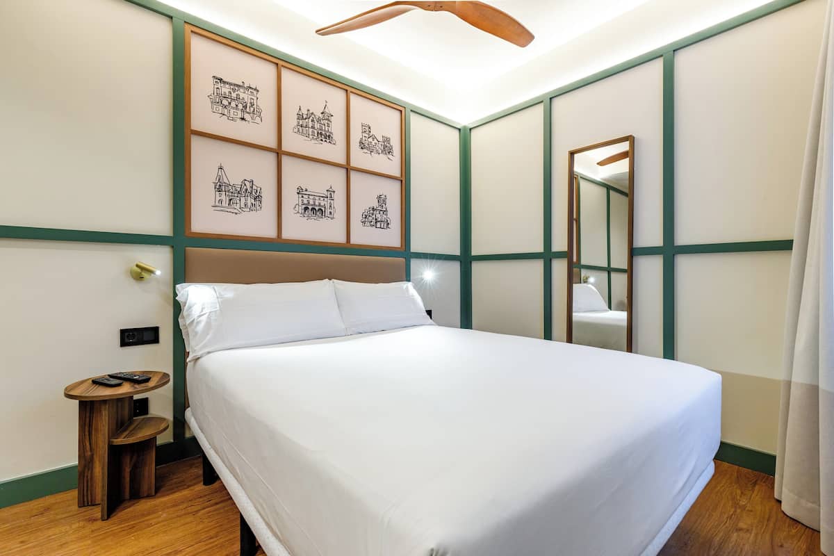 A neatly made bed in a hotel room with a headboard, a small wooden side table with a remote control, and a wall-mounted mirror.