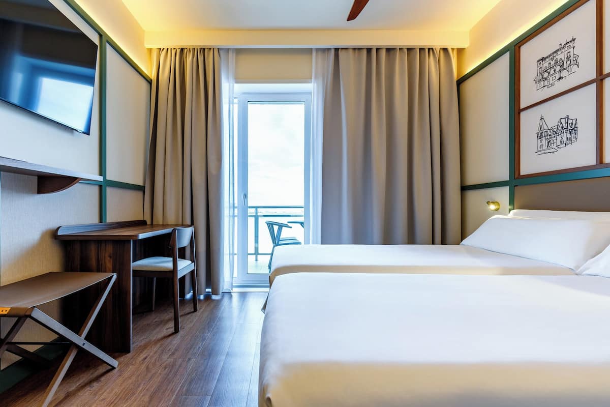 A hotel room with a bed, a desk, a chair, a TV, and a balcony with curtains.