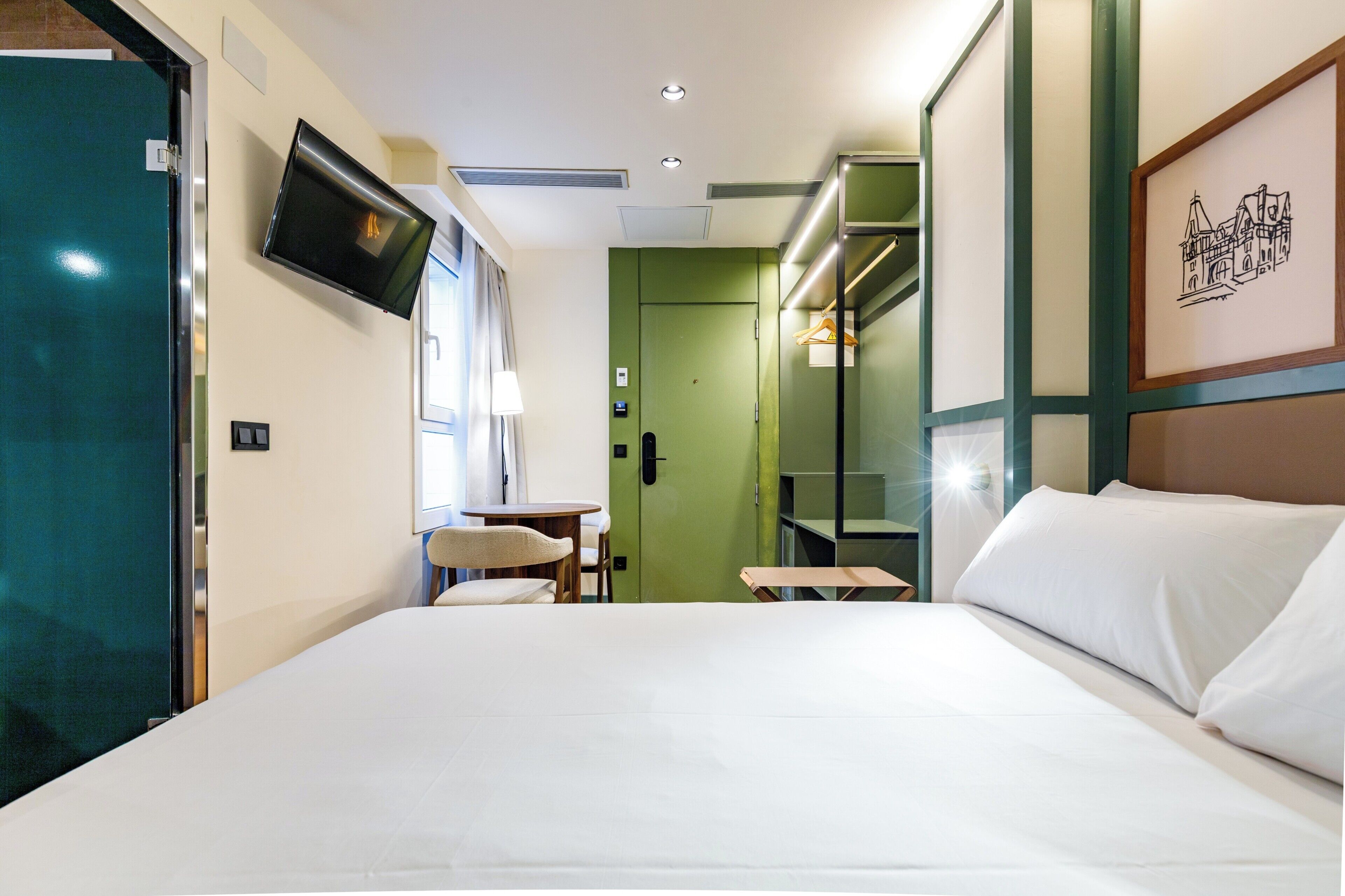 Habitación de hotel con cama, televisor montado en la pared, una mesita con una silla y una puerta verde.