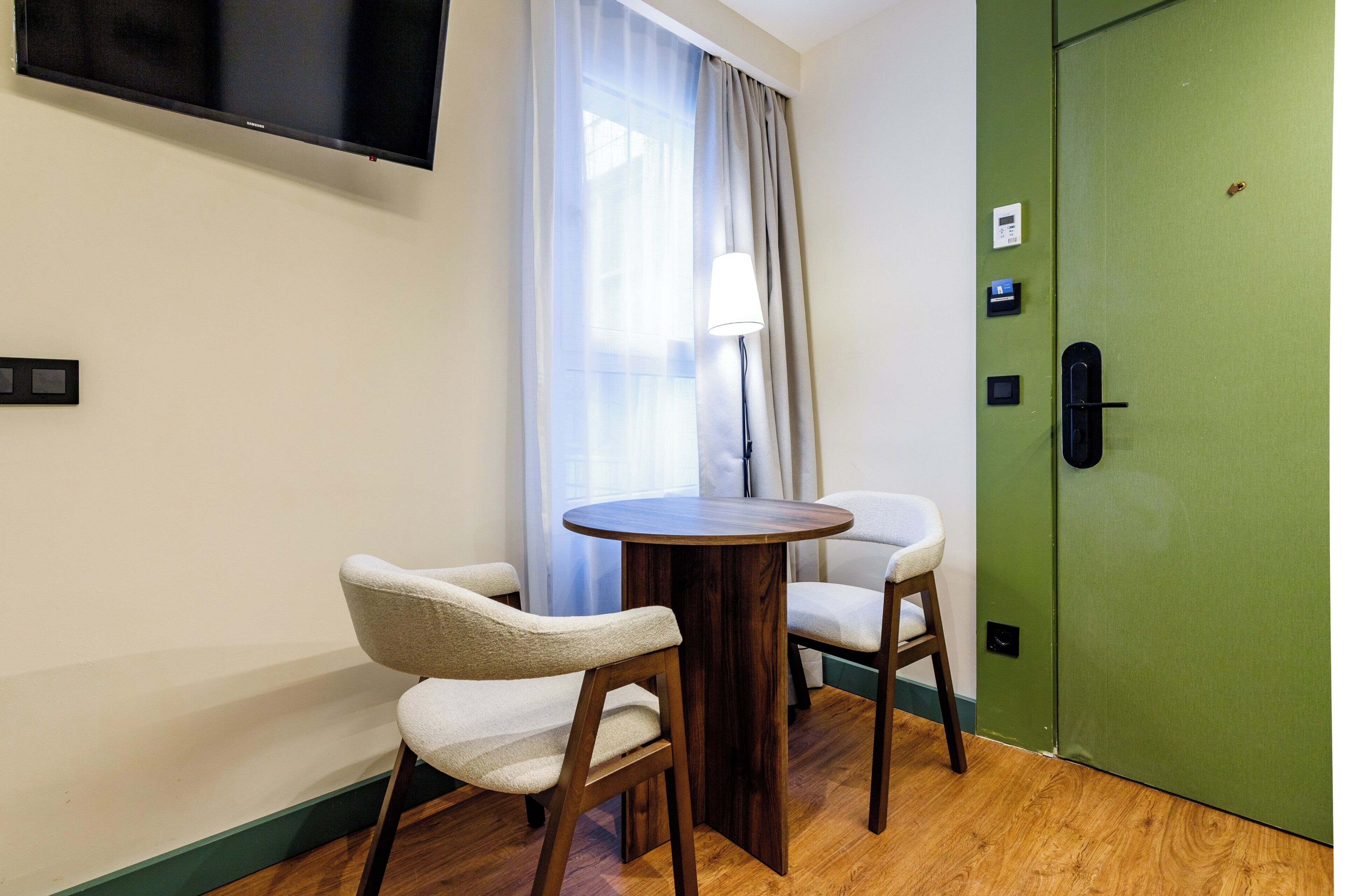 Una habitación con una mesa de madera, dos sillas, un televisor montado en la pared, una puerta verde y una ventana con cortinas.