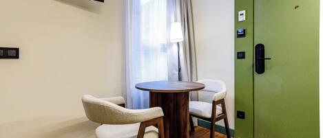 Una habitación con una mesa de madera, dos sillas, un televisor montado en la pared, una puerta verde y una ventana con cortinas.
