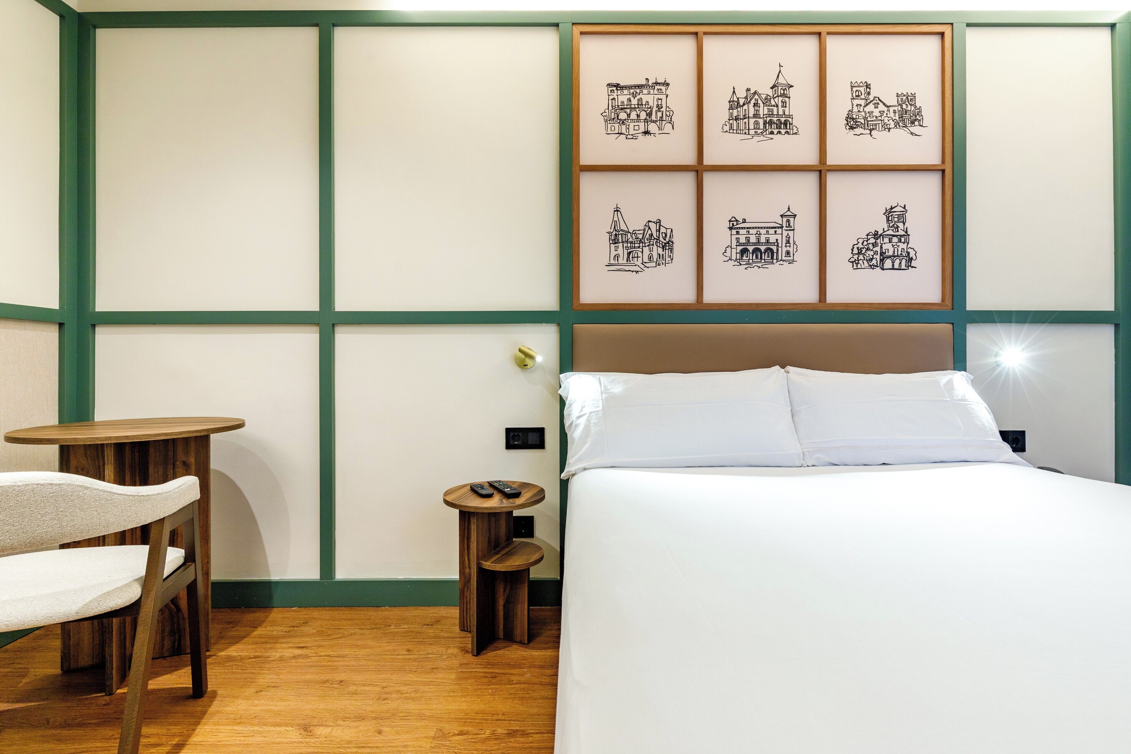 A hotel room with a bed, a small round table with a chair, a bedside table with a lamp, and a wall with framed pictures.