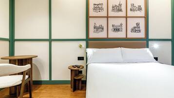 A hotel room with a bed, a small round table with a chair, a bedside table with a lamp, and a wall with framed pictures.