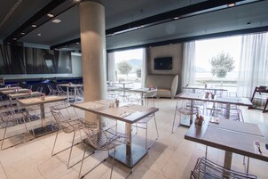 A modern dining area with multiple tables and chairs, large windows with a view of the outdoors, and a flat-screen TV mounted on the wall.