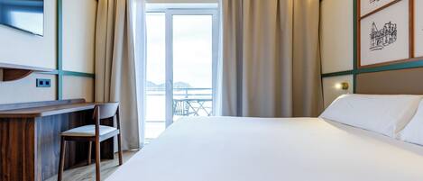 A hotel room with a bed, a desk with a chair, a TV, and a balcony with curtains.