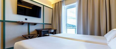 A hotel room with a bed, a desk, a chair, a flat-screen TV mounted on the wall, and a window with curtains.