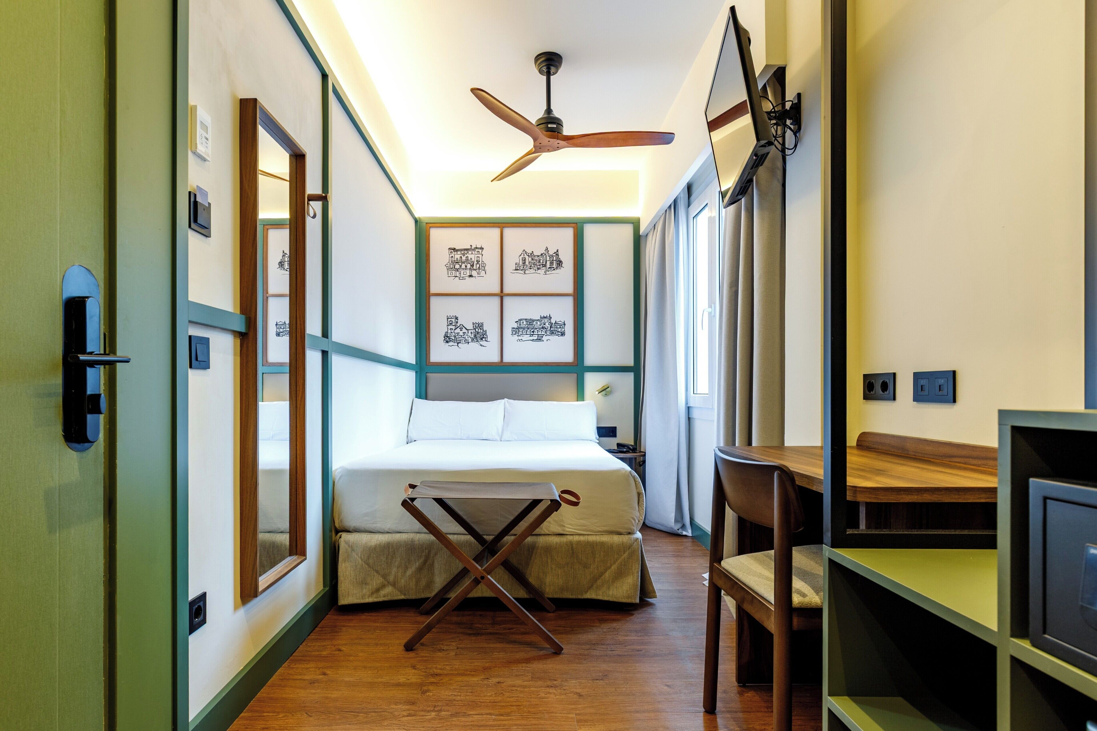 Ein schmales Hotelzimmer mit einem Bett, einem Schreibtisch, einem Stuhl, einem Deckenventilator und gerahmten Bildern an der Wand.