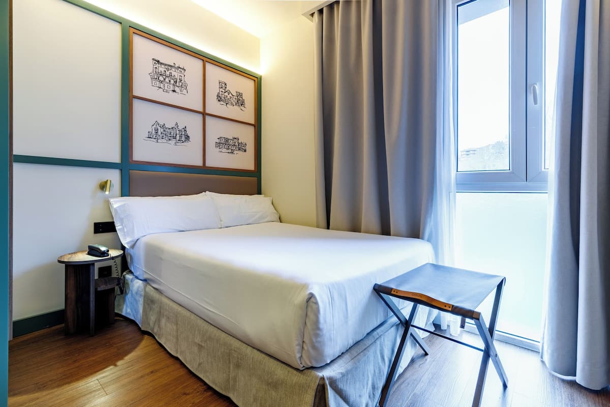 A hotel room with a bed, a small table, a nightstand, and a window with curtains.