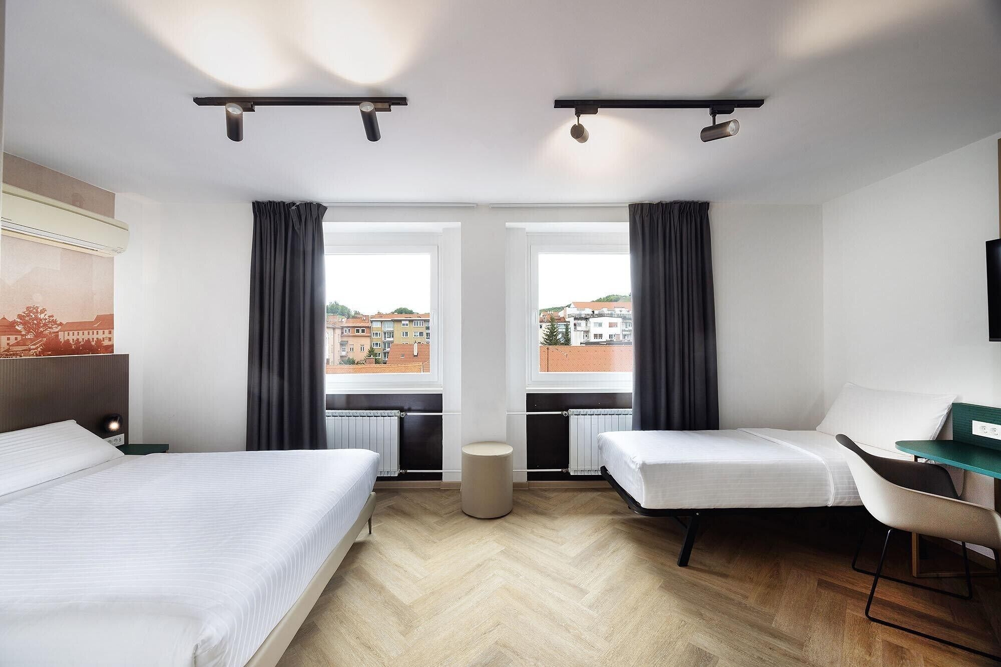 Et hotelværelse med to senge, et skrivebord og udsigt til bygninger gennem store vinduer.
