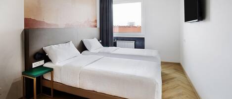 A hotel room with two beds, a small table, a wall-mounted TV, and a window with a view of buildings.