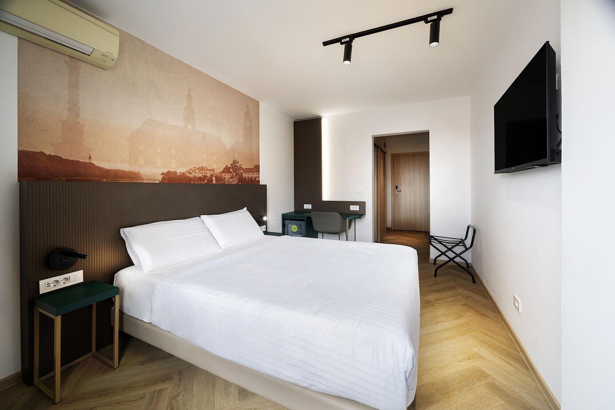 A hotel room with a large bed, a desk, a chair, a flat-screen TV, and a wall-mounted air conditioner.