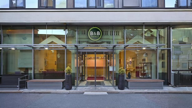 Un hotel B&B con fachada de vidrio moderna, un letrero verde y una entrada acogedora con bancos y plantas en macetas.