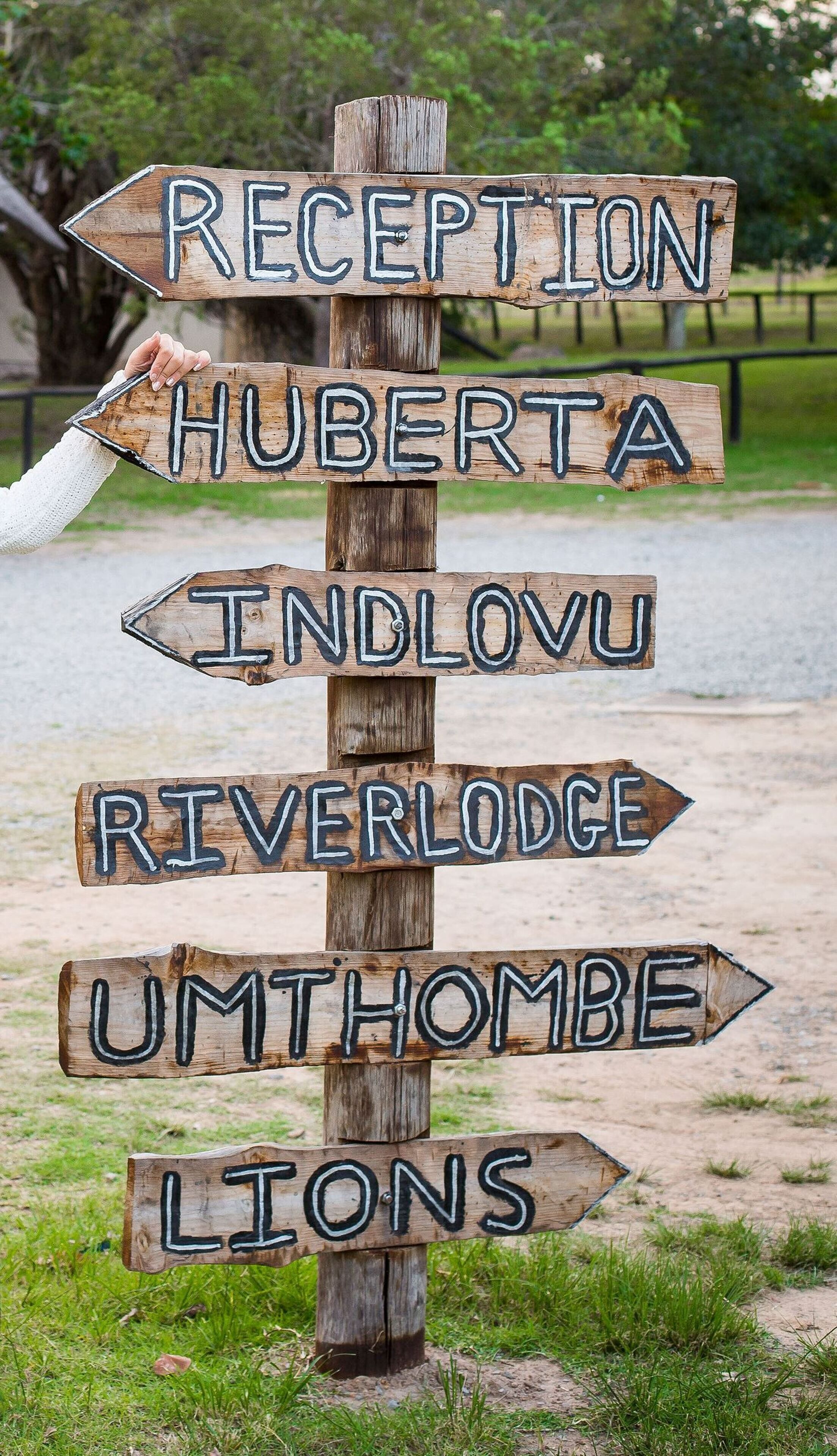 A wooden signpost with directional arrows pointing to various locations such as Reception, Huberta, Indlovu, Riverlodge, Umthombe, and Lions.